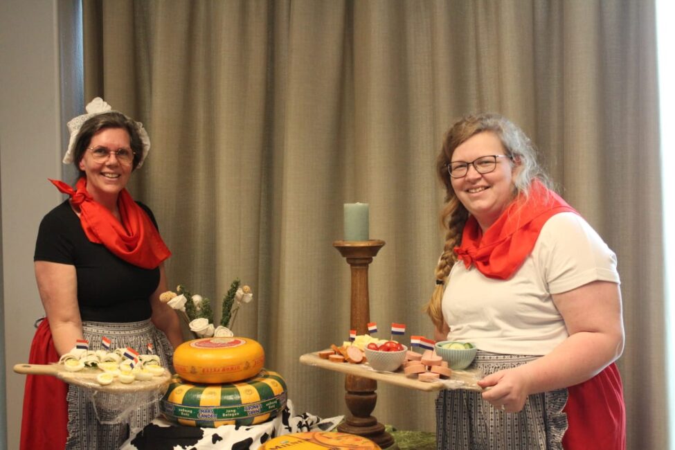 Twee vrouwen in traditionele kleding presenteren kaas en hapjes op houten planken, met een kaars en gordijn op de achtergrond.