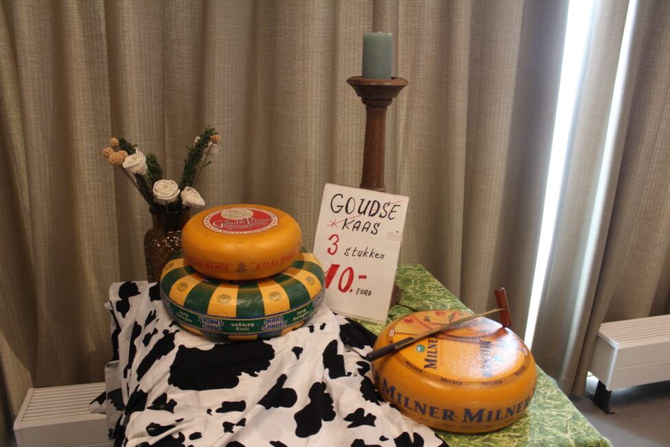 Een tafel met drie grote kazen, een kaars op een houder, en een bord met de tekst "Goudse kaas 3 stuks 10 euro".