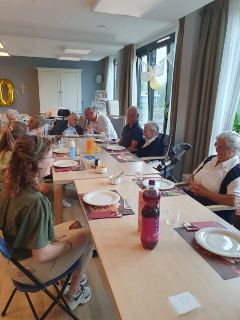 Een groep mensen van verschillende leeftijden zit aan een lange tafel in een lichte kamer, klaar voor een maaltijd.