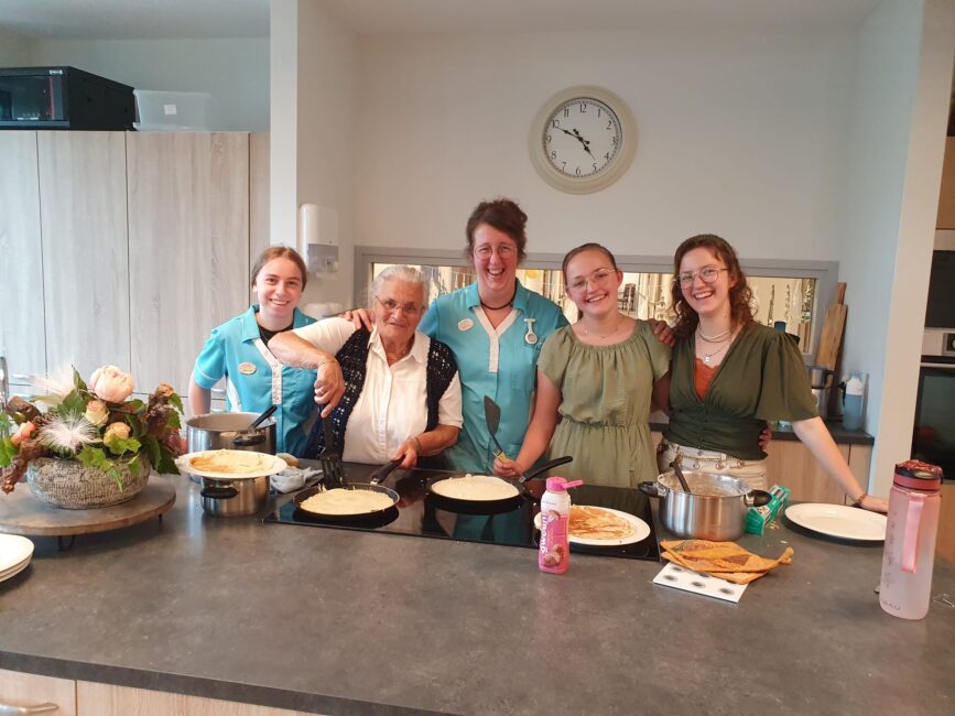 Vijf vrouwen staan lachend in een keuken, met pannenkoeken op het aanrecht. Er hangt een klok aan de muur.
