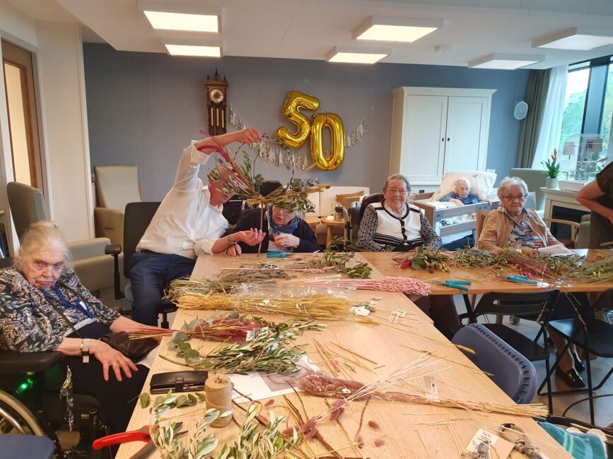 Een groep ouderen maakt bloemstukken aan een tafel. Op de achtergrond hangt een gouden "50" ballon aan de muur.
