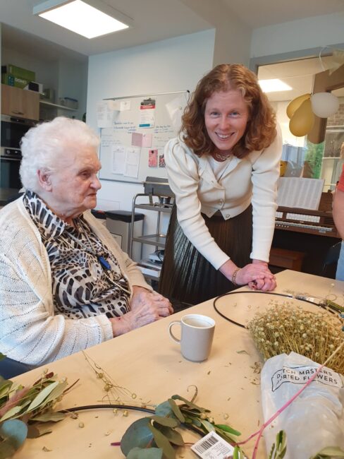 Twee vrouwen in een kamer, een oudere zittend aan tafel met bloemen, de ander staat glimlachend naast haar.