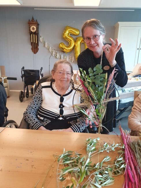 Twee vrouwen glimlachen terwijl ze een boeket bloemen vasthouden. Op de achtergrond hangt een gouden "50" ballon.