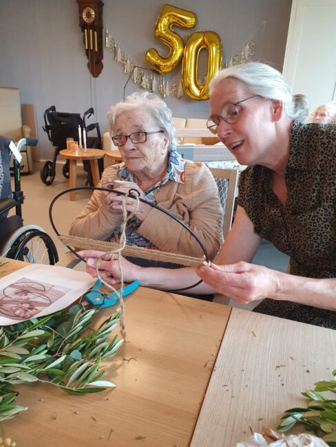 Twee vrouwen maken een bloemstuk aan tafel. Op de achtergrond zijn gouden ballonnen in de vorm van het getal 50 te zien.