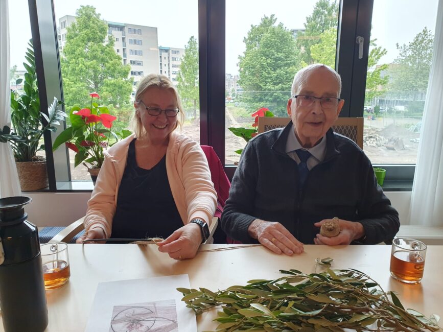 Een oudere man en vrouw zitten aan een tafel met takken en thee, omringd door planten, voor een raam met uitzicht op bomen.