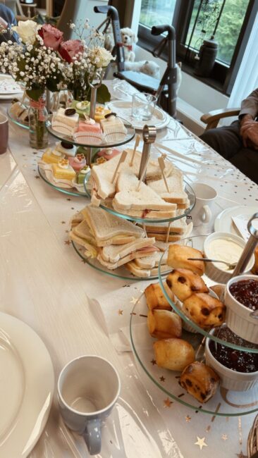 Een gedekte tafel met sandwiches, gebakjes en jam. Er staan bloemen in een vaas en een rolstoel is op de achtergrond zichtbaar.