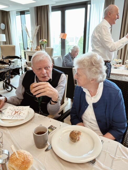 Oudere man en vrouw zitten aan tafel in een eetzaal, pratend en genietend van een maaltijd met broodjes en koffie.