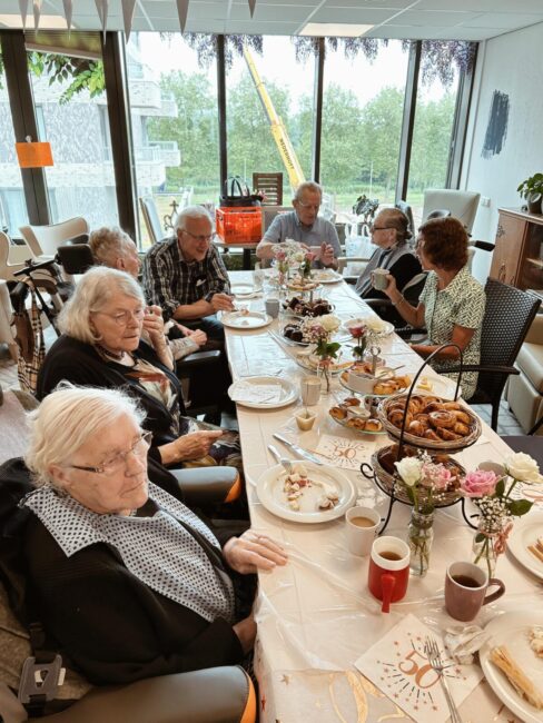 Groep ouderen zit aan een feestelijk gedekte tafel in een lichte ruimte, genietend van een maaltijd en elkaars gezelschap.