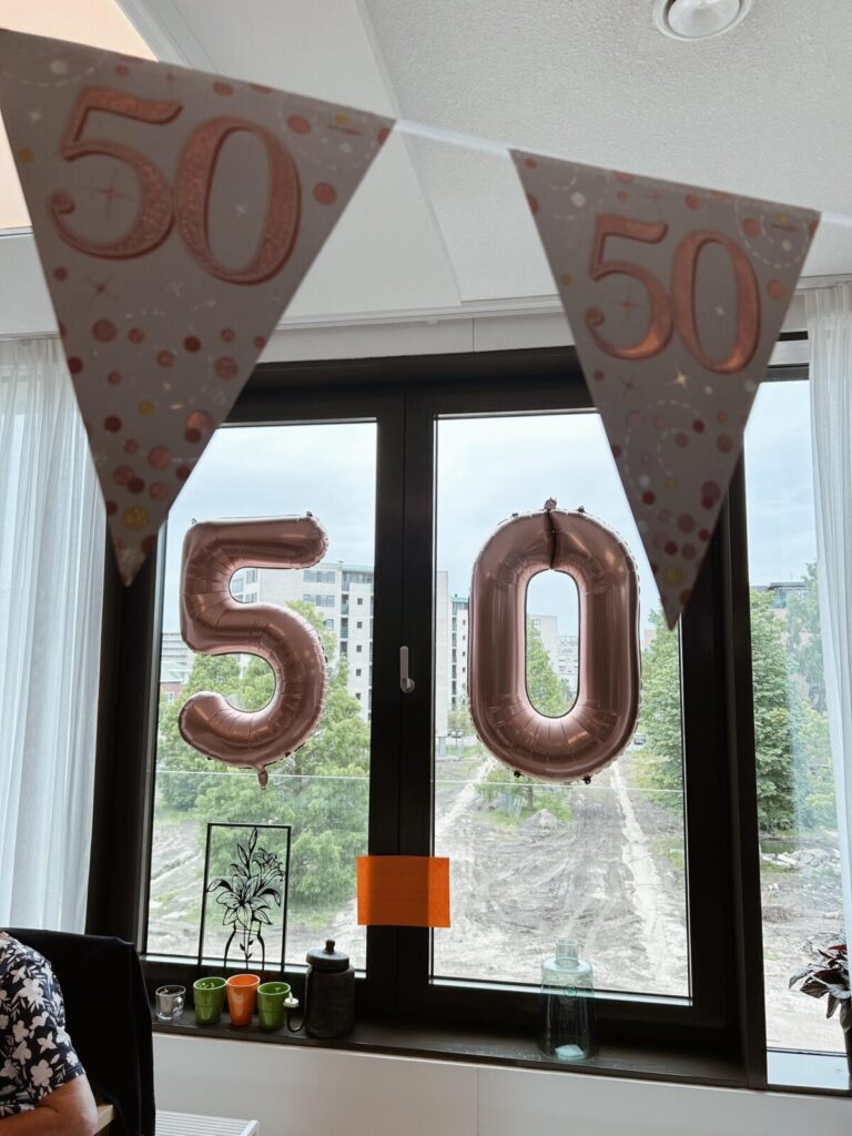 Vlaggen en ballonnen met het getal 50 hangen voor een raam, ter viering van een 50e verjaardag in een kamer.