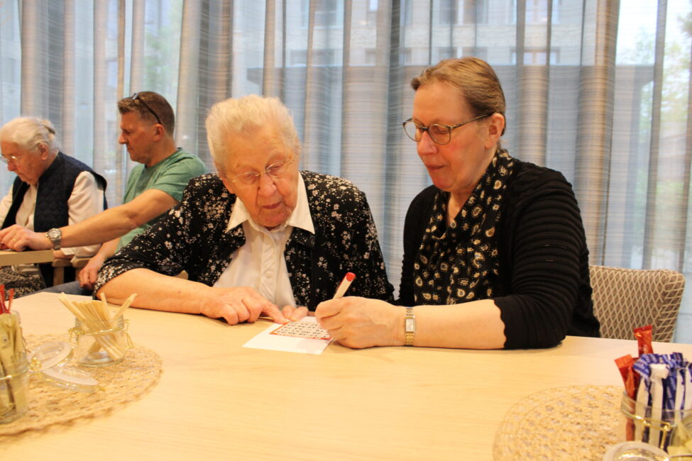 Twee oudere vrouwen zitten aan een tafel en werken samen aan een puzzel. Op de achtergrond zijn meer mensen zichtbaar.