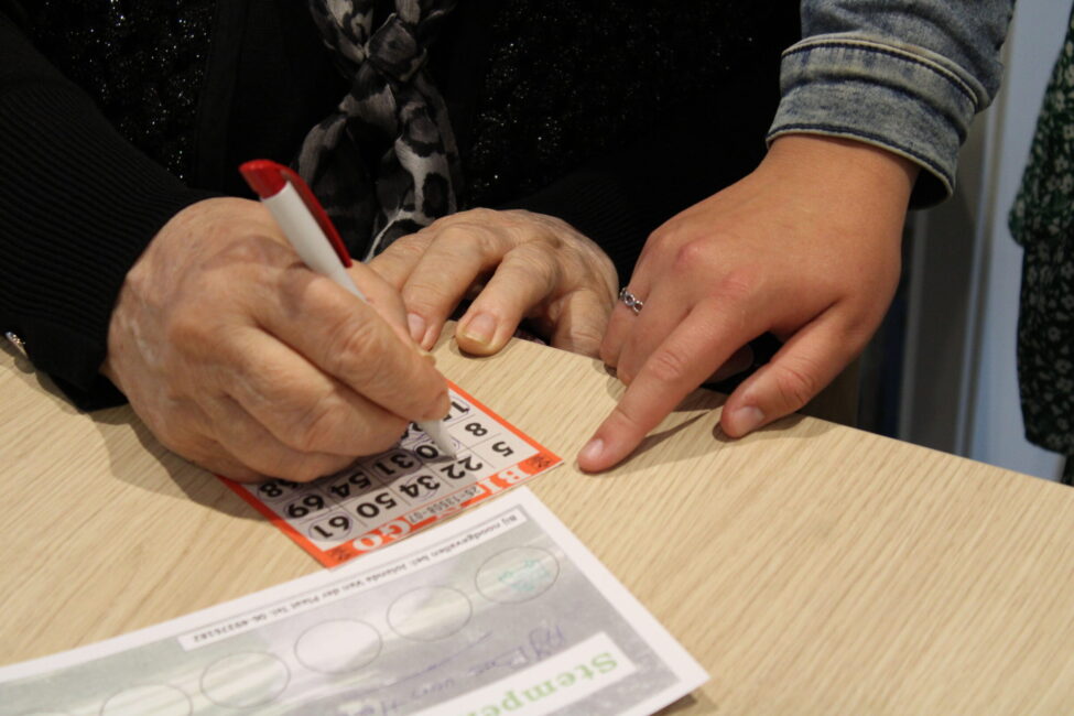 Twee personen spelen bingo; een oudere hand markeert een kaart met een rode stift, terwijl een jongere hand aanwijzingen geeft.