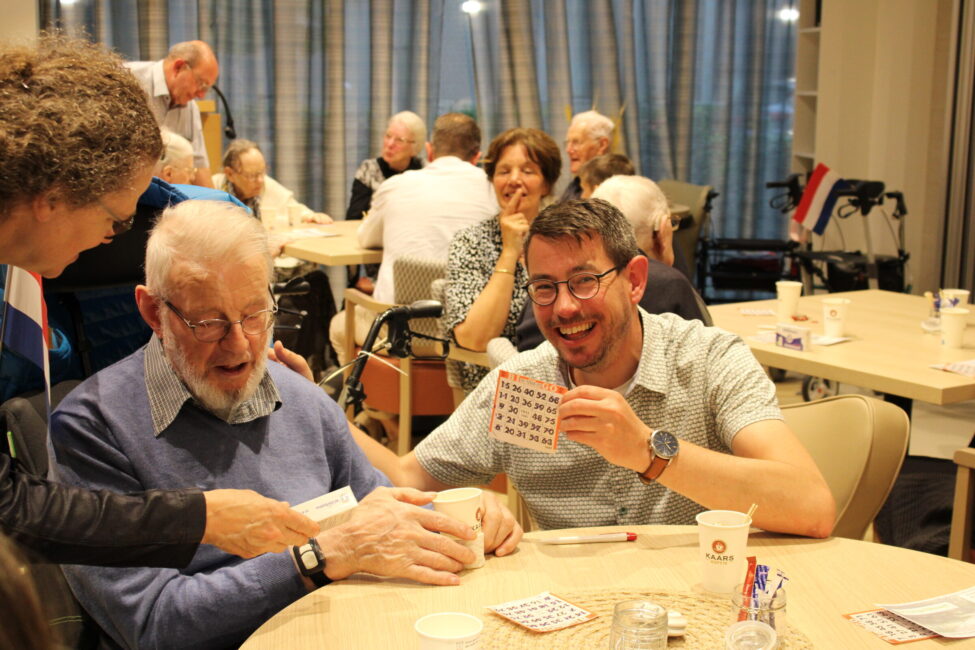 Een groep mensen speelt bingo in een gezellige ruimte. Een oudere man en een jongere man lachen terwijl ze hun kaarten bekijken.