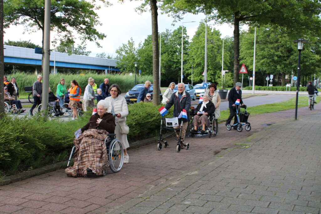 Een groep mensen duwt rolstoelen met ouderen over een trottoir, omringd door groen en bomen, met Nederlandse vlaggetjes.
