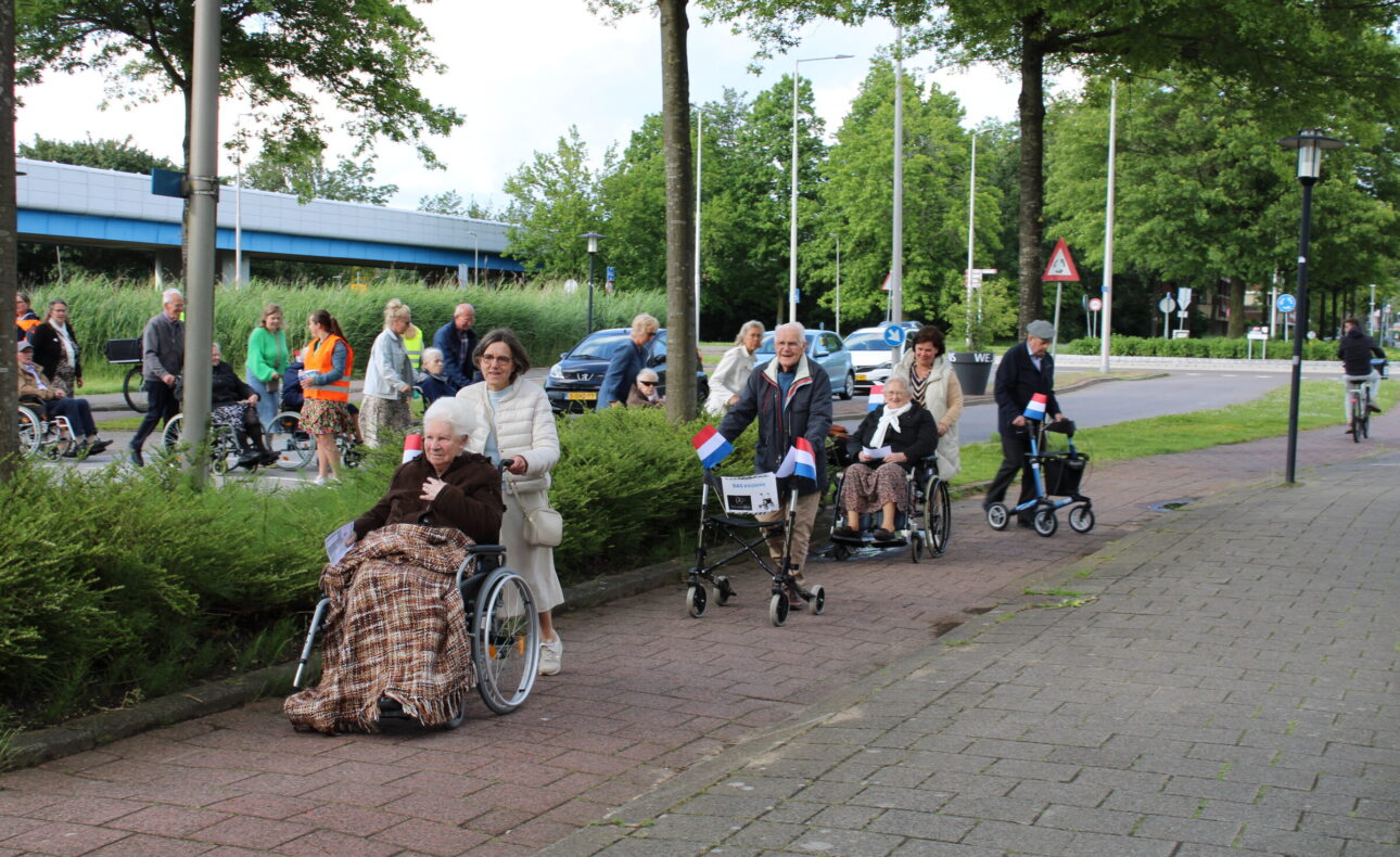 Een groep mensen duwt rolstoelen met ouderen over een trottoir, omringd door groen en bomen, met Nederlandse vlaggetjes.