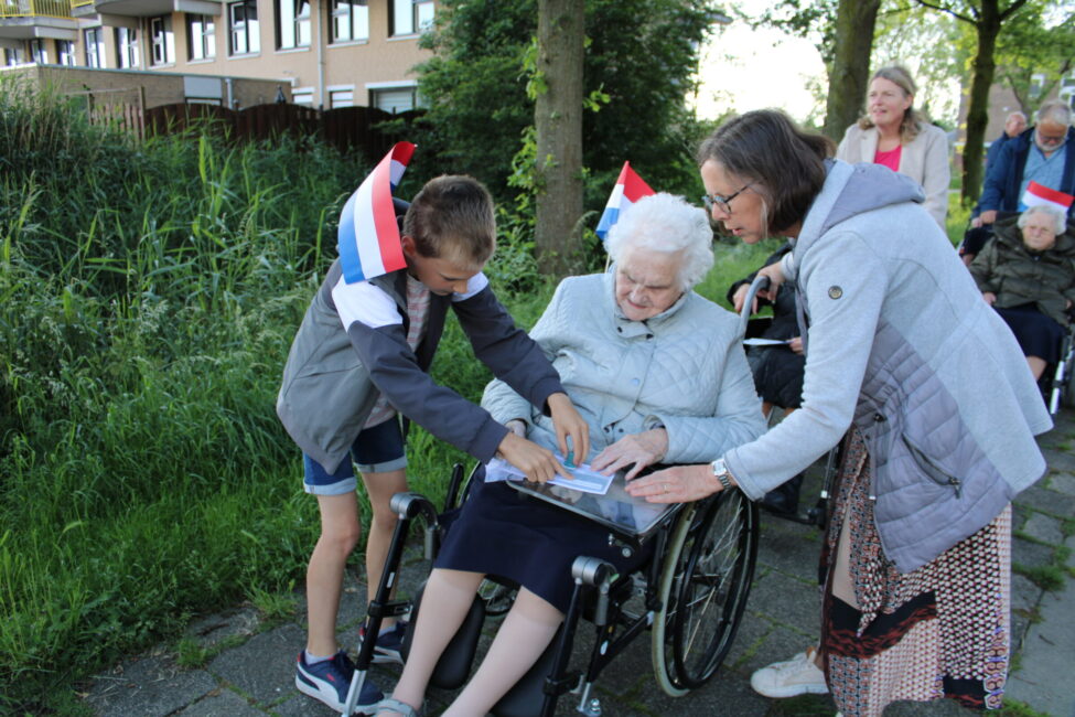 Een jongen en een vrouw helpen een oudere dame in een rolstoel met een boek. Ze dragen feestelijke hoedjes buiten.