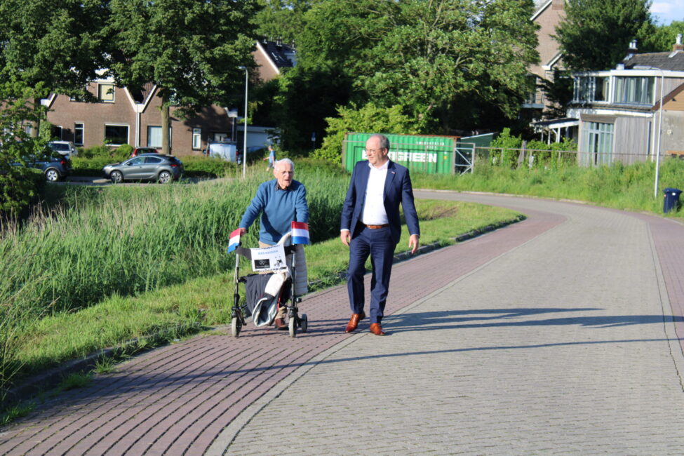 Twee mannen wandelen op een zonnige dag. De oudere man gebruikt een rollator, terwijl de jongere man naast hem loopt.