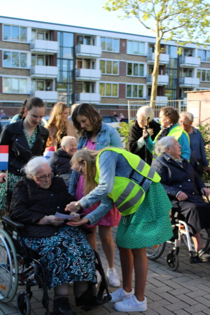 Groep mensen, waaronder ouderen in rolstoelen en jongere begeleiders met gele hesjes, buiten voor een gebouw.