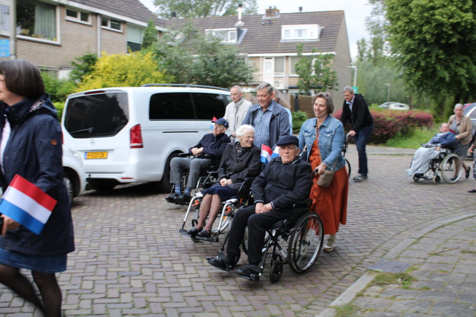 Mensen duwen rolstoelen met ouderen tijdens een buitenactiviteit. Sommigen dragen Nederlandse vlaggen. Auto's op de achtergrond.