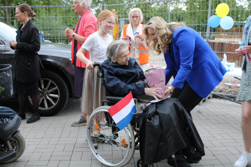 Een vrouw in een rolstoel met een Nederlandse vlag krijgt een boeket bloemen van een andere vrouw in een blauwe jas.