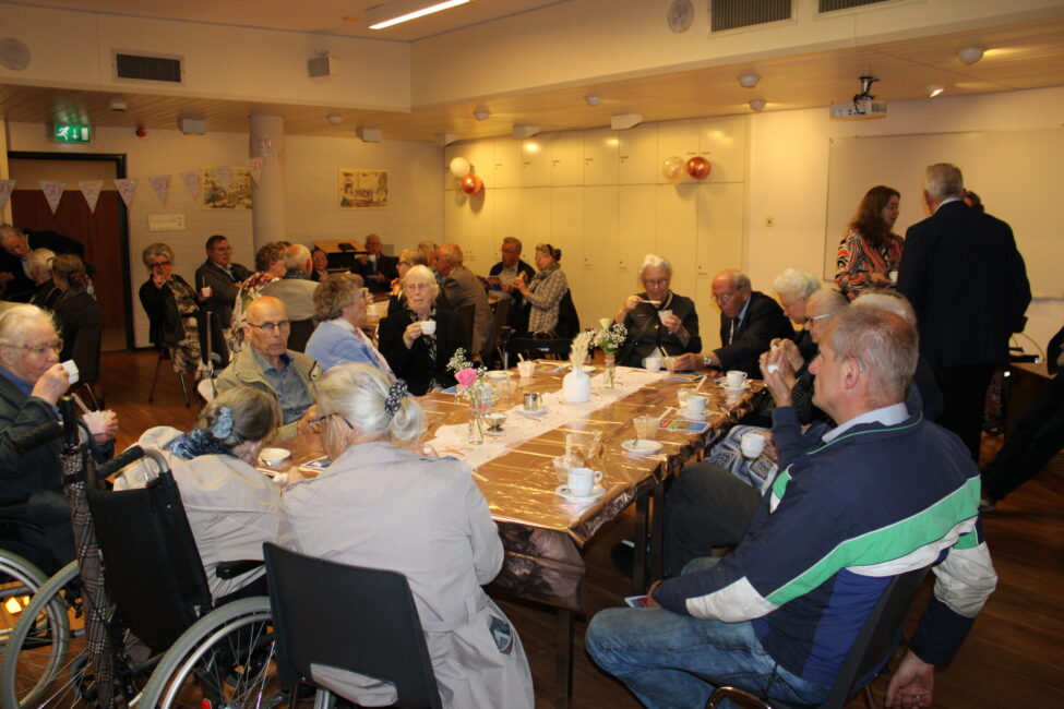 Groep ouderen zit aan tafels in een zaal, genietend van koffie en gesprek. De sfeer is gezellig en ontspannen.