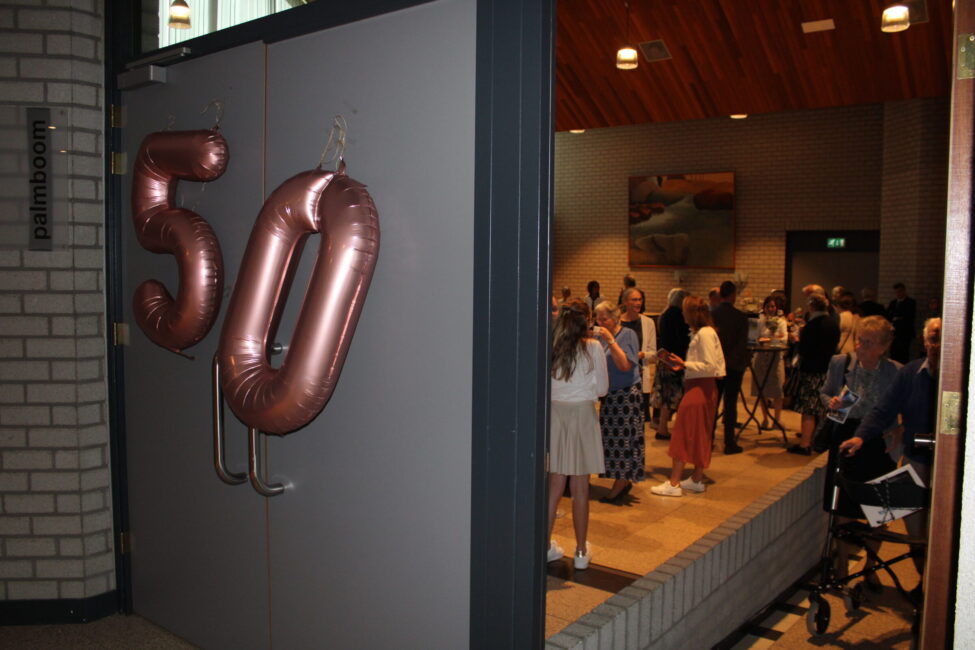 Een feestelijke ruimte met een open deur versierd met ballonnen in de vorm van het getal 50. Mensen staan binnen te praten.