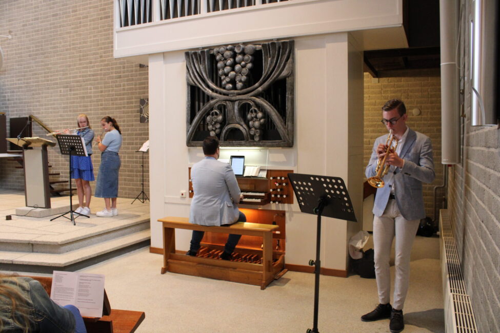 Muzikanten spelen in een kerk: een organist, een trompettist en twee zangeressen. Ze staan voor een modern kunstwerk.