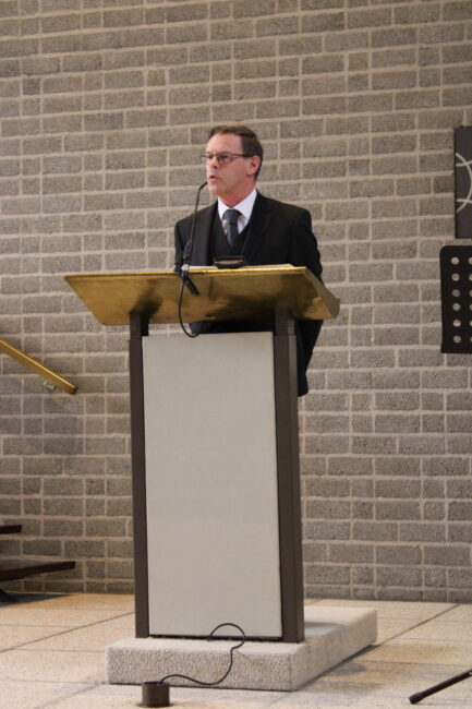 Een man in een pak spreekt achter een lessenaar in een zaal met bakstenen muren. Er is een microfoon op de lessenaar.