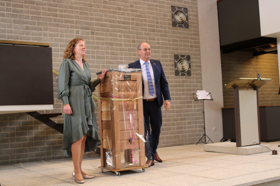 Twee mensen staan naast een stapel dozen op een karretje in een zaal met een grijze bakstenen muur en een lessenaar.