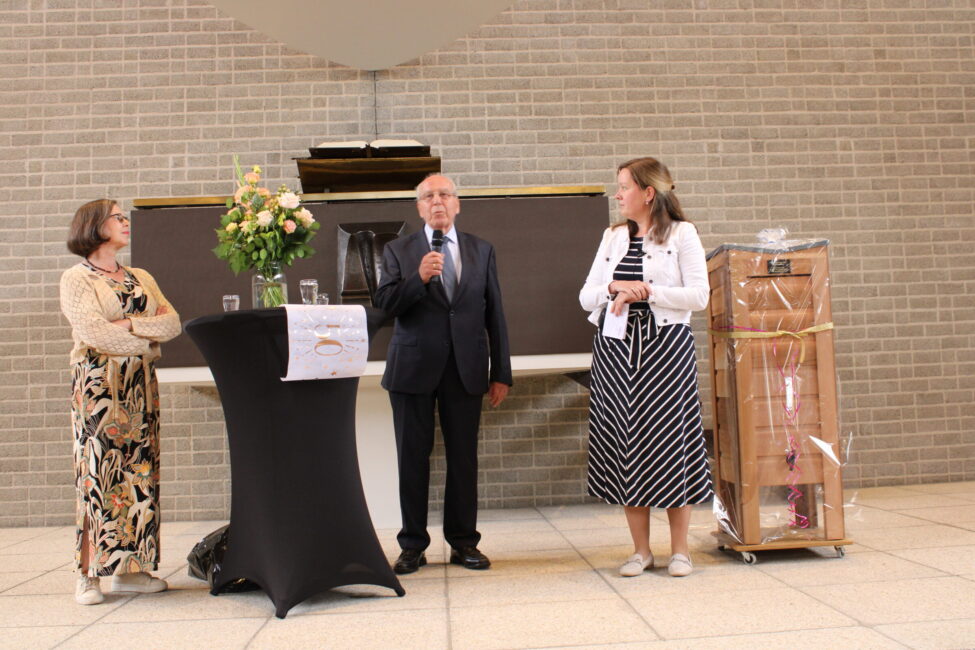Drie mensen staan bij een tafel met bloemen en een microfoon in een zaal met bakstenen muur. Er is een ingepakt cadeau.