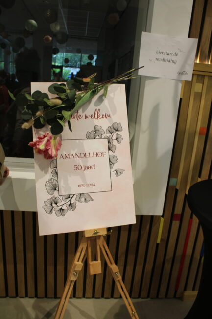 Een bord op een ezel met bloemen en de tekst "Amandelhof 30 jaar". Ernaast hangt een bordje met "Welkom in Amandelhof".