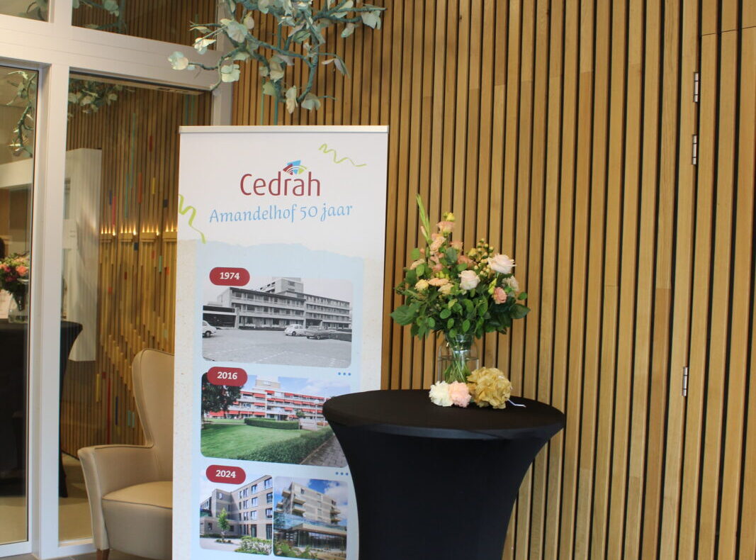 Een banner van Cedrah naast een hoge tafel met bloemen, geplaatst in een moderne ruimte met houten wandbekleding.
