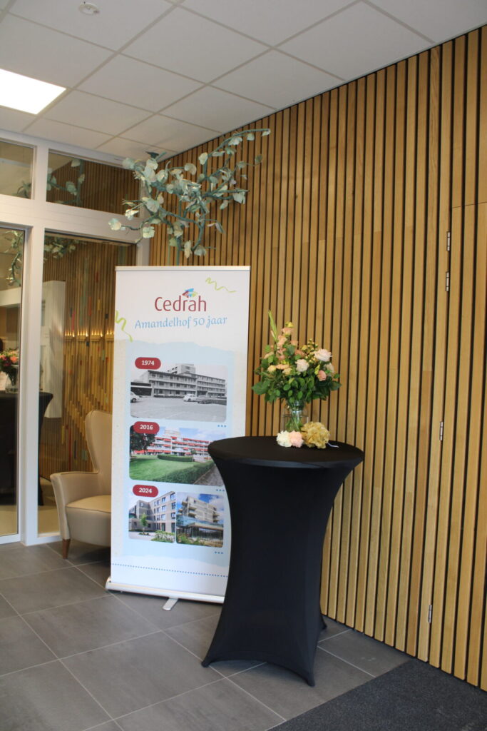 Een banner van Cedrah naast een hoge tafel met bloemen, geplaatst in een moderne ruimte met houten wandbekleding.