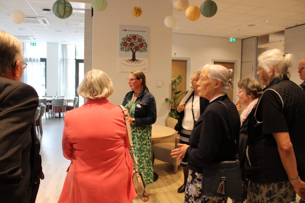 Groep oudere mensen in gesprek met een vrouw in een groene jurk in een gemeenschappelijke ruimte met decoratieve ballen.