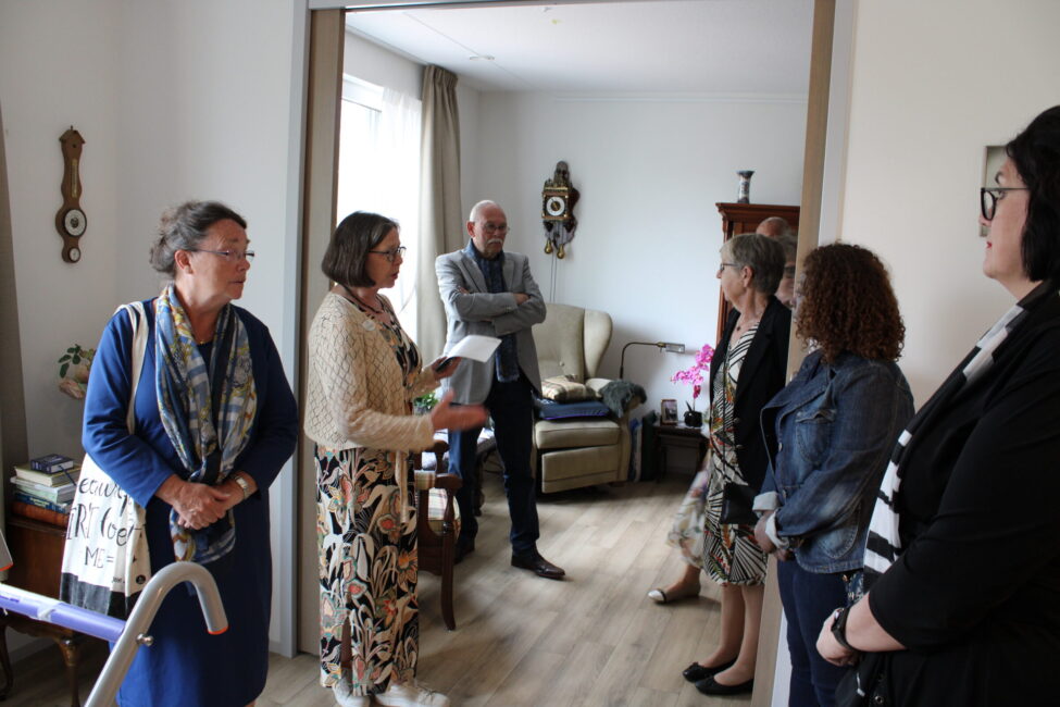 Groep mensen staat in een woonkamer en praat met elkaar. Er zijn meubels en decoraties zichtbaar in de achtergrond.
