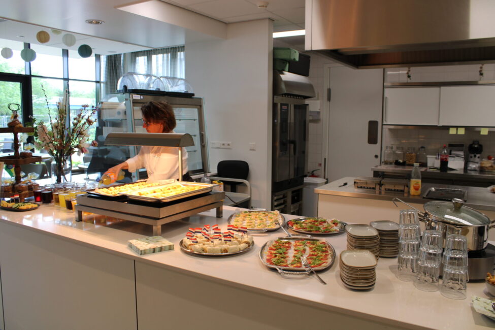 Een moderne keuken met een kok die gerechten bereidt. Op de voorgrond staan schalen met diverse hapjes en glazen.