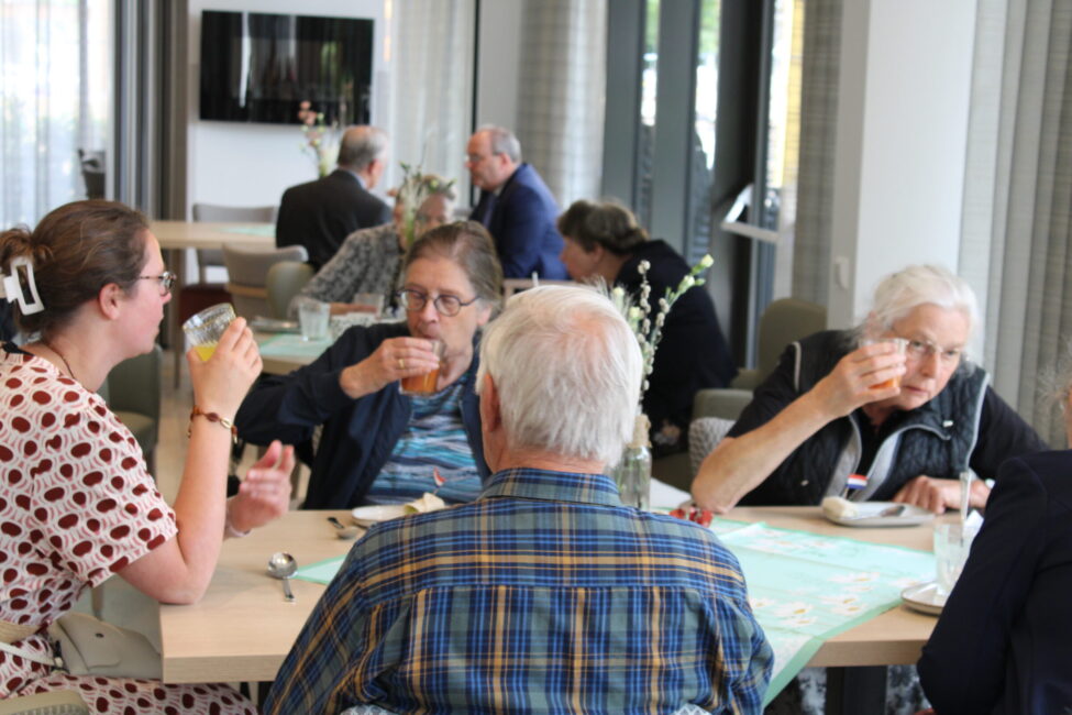 Groep mensen zit aan een tafel in een eetzaal, genietend van drankjes en gesprekken. De sfeer is ontspannen en gezellig.