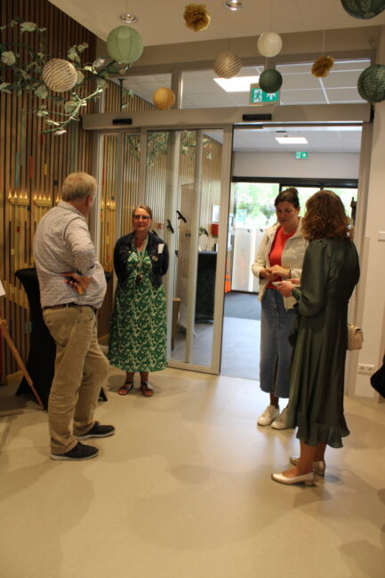 Groep mensen in gesprek in een moderne ruimte met glazen deuren en decoraties aan het plafond. Ze dragen casual kleding.