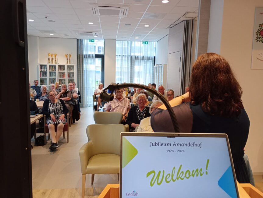 Een vrouw spreekt voor een groep mensen in een zaal. Op de achtergrond is een scherm met de tekst "Jubileum Amandelhof".