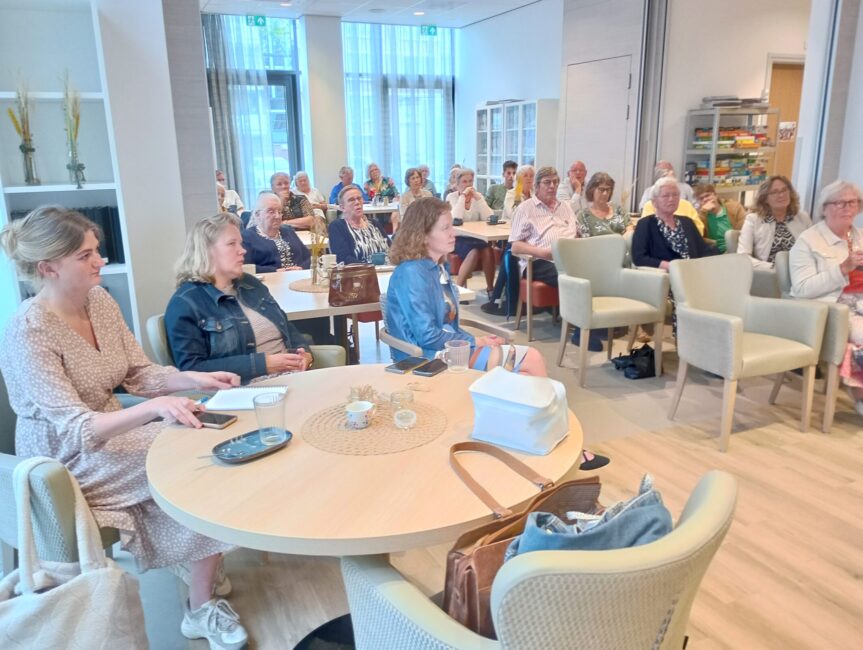 Groep mensen zit in een lichte ruimte met tafels en stoelen, aandachtig luisterend naar een presentatie of spreker.
