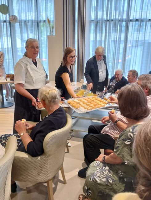 Groep mensen in een lichte ruimte, zittend en staand, terwijl een vrouw hapjes serveert van een schaal met oranje snacks.