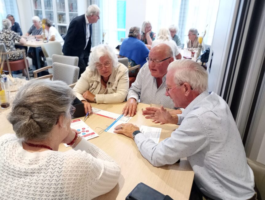 Een groep oudere mensen zit aan een tafel in een lichte ruimte, verdiept in een gesprek. Op tafel liggen papieren.