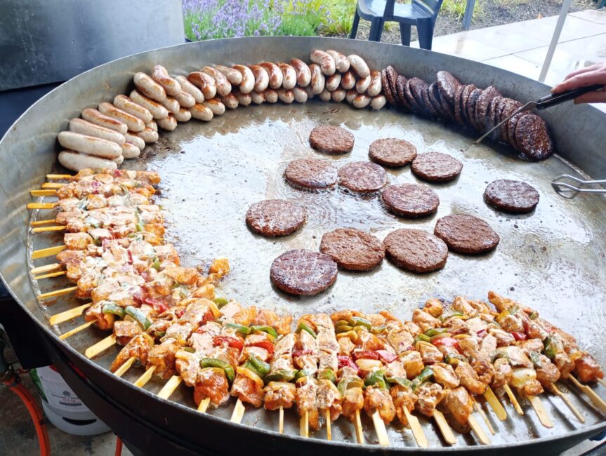 Grote pan met diverse soorten vlees op de barbecue: spiesjes, hamburgers en worsten, klaar om gegrild te worden.