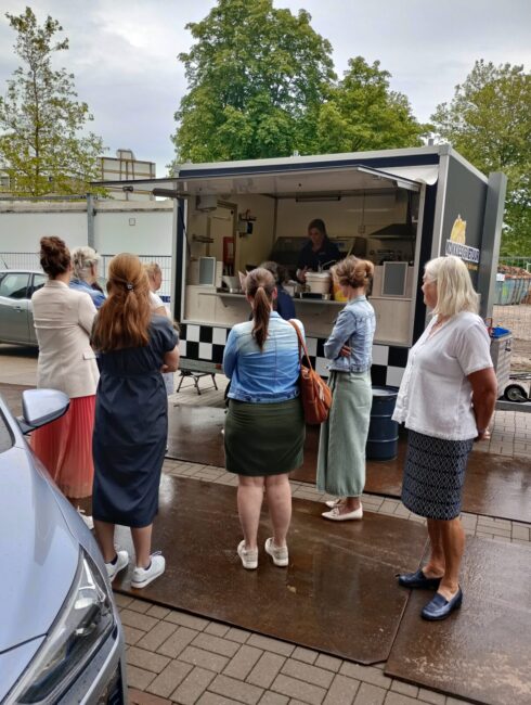 Groep mensen staat buiten bij een foodtruck op een regenachtige dag. Ze wachten op hun bestelling. Bomen op de achtergrond.