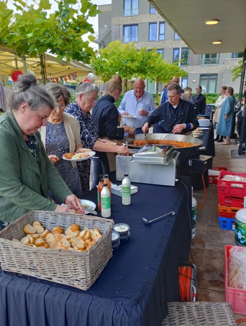 Groep mensen geniet van een buffet buiten, met broodjes en sauzen op tafel. Op de achtergrond zijn bomen en een gebouw te zien.