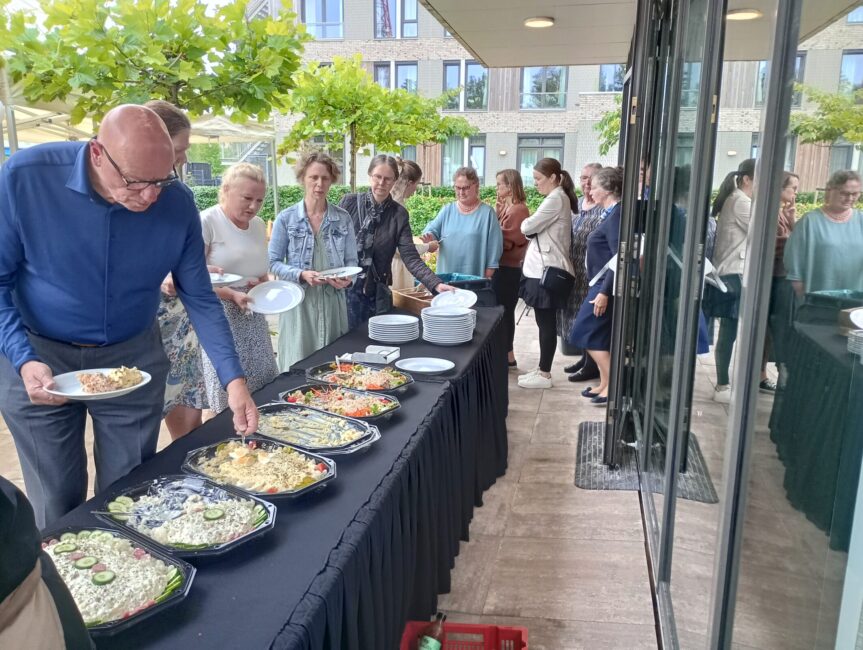 Groep mensen staat in de rij bij een buffet met diverse gerechten op een tafel buiten, naast een gebouw met grote ramen.
