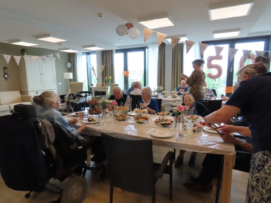 Een groep mensen zit aan een feestelijk gedekte tafel in een kamer met slingers en een groot raam. Er hangt een "50" decoratie.