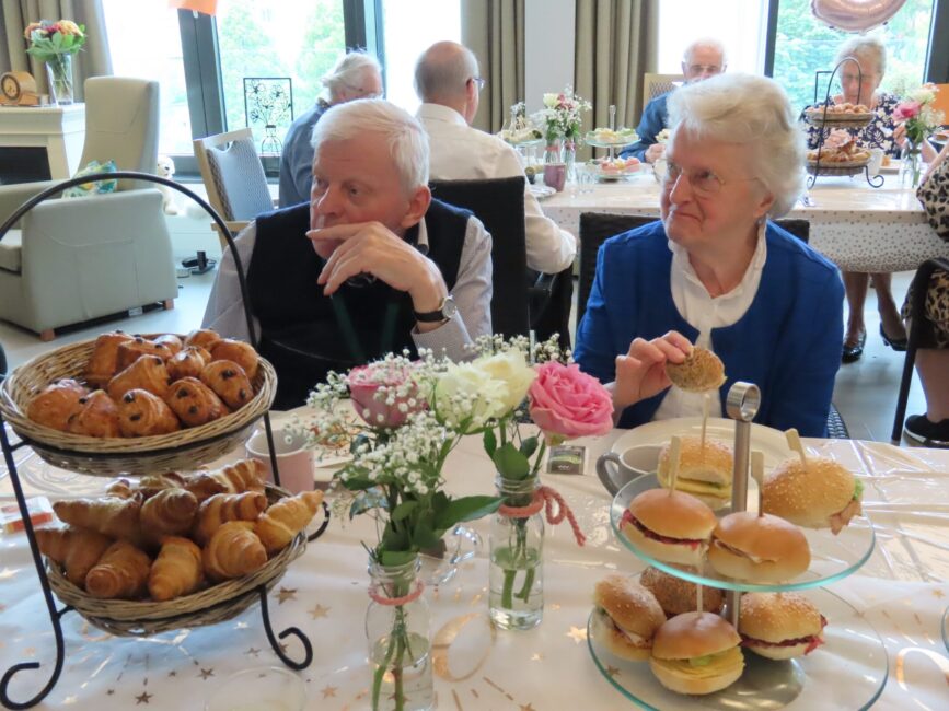 Twee oudere mensen zitten aan een tafel met diverse broodjes en bloemen, in een gezellige ruimte met andere mensen op de achtergrond.