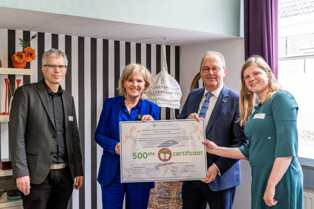 Vier mensen poseren met een groot certificaat in een kamer met gestreepte muur en raam. Een vrouw draagt een blauw pak.