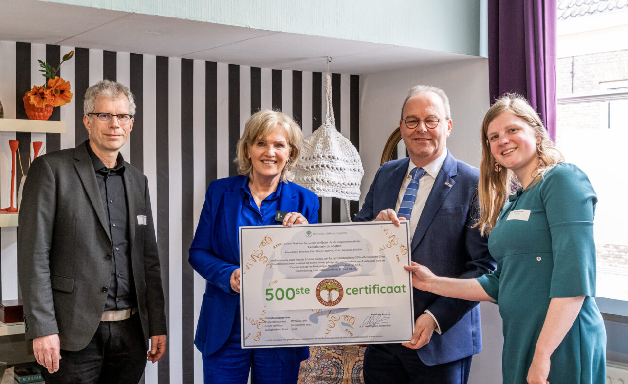 Vier mensen poseren met een groot certificaat in een kamer met gestreepte muur en raam. Een vrouw draagt een blauw pak.