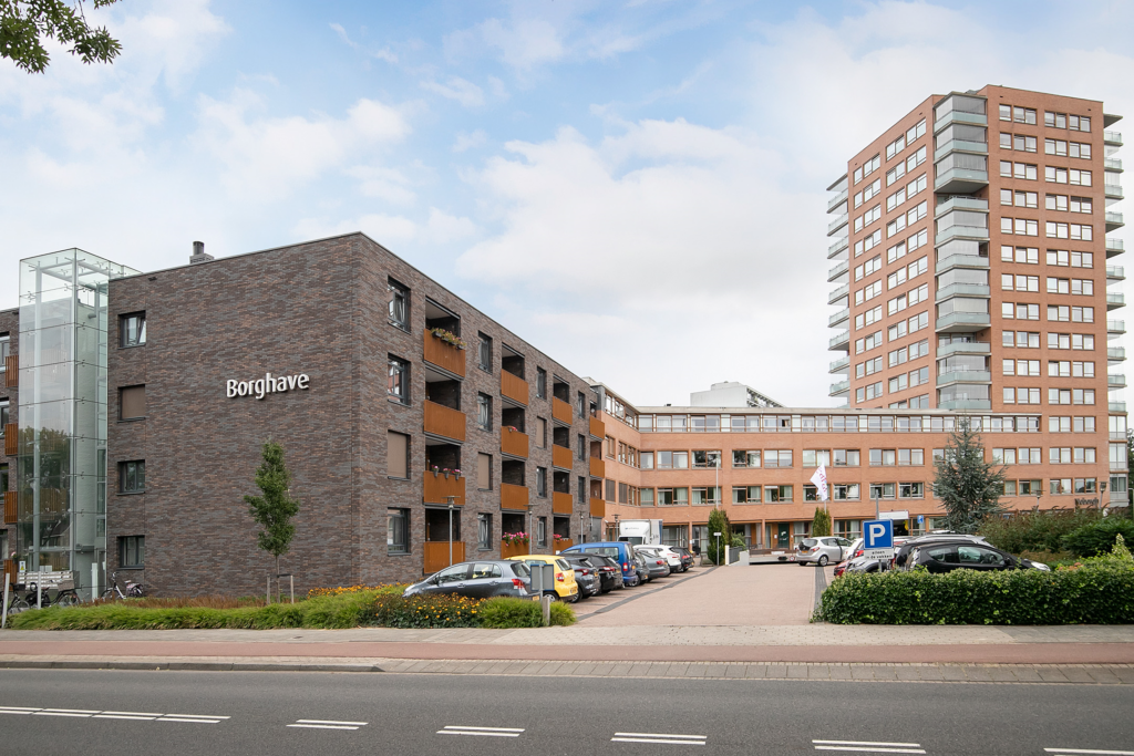 Afbeelding van een modern appartementencomplex met meerdere verdiepingen, geparkeerde auto's en een blauwe lucht op de achtergrond.